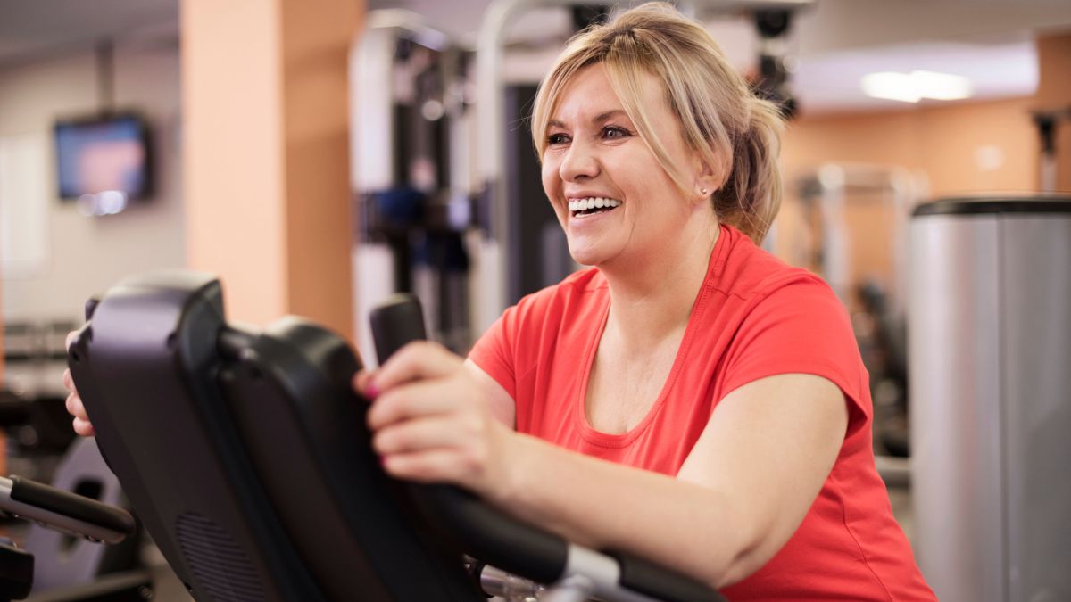 A woman is shown smiling and pedaling on a workout bicycle.